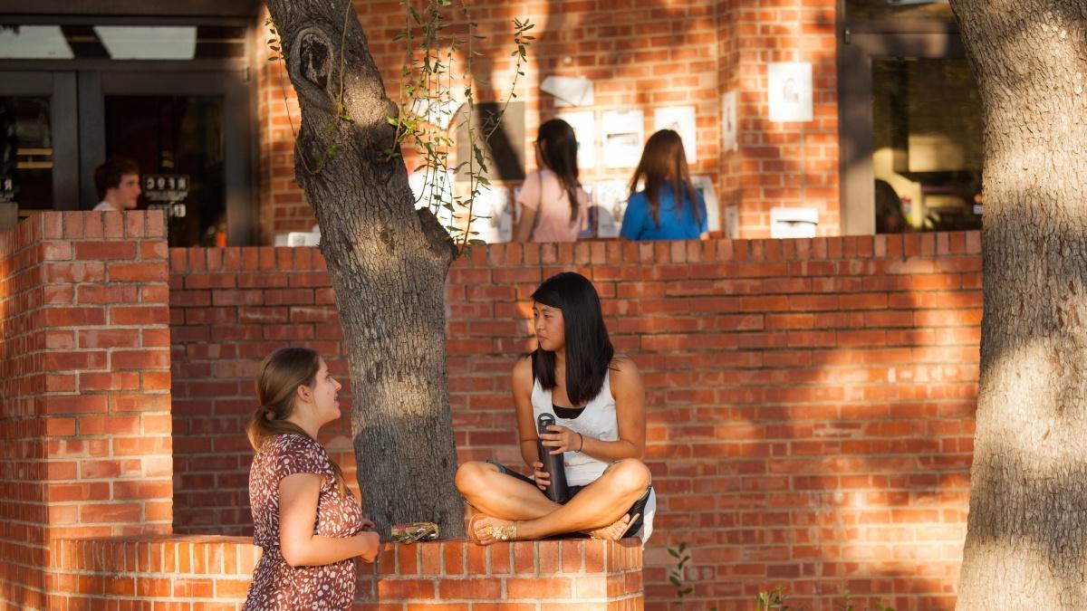 两个学生坐在梅比食堂前的红砖墙上